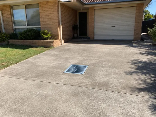 Concrete driveway with a grated outdoor drain beside a brick house and garage in Swansea Heads