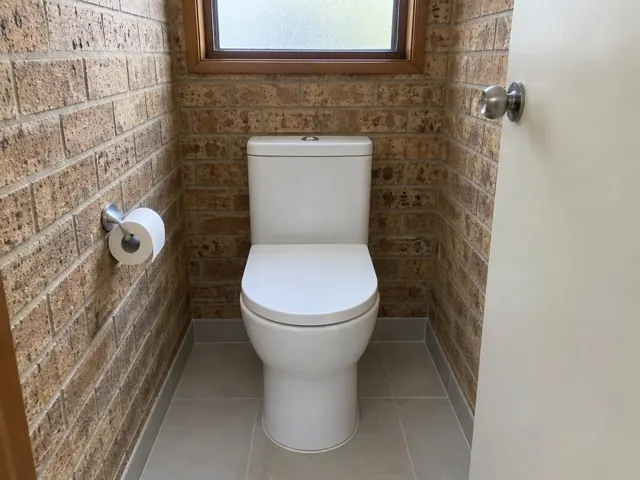 White toilet in a small brick-walled bathroom with tiled floor and frosted window in Caves Beach